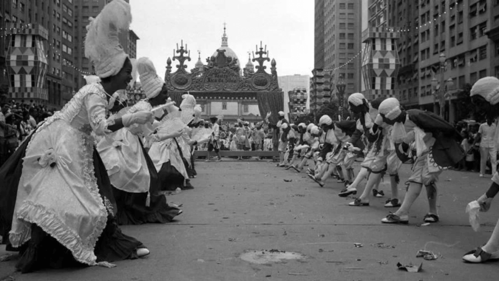 Black and white image of a cultural parade with participants in elaborate costumes, including large headpieces and formal attire, dancing in the streets.