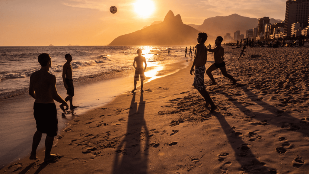 Golden sunset at Rio de Janeiro beach with people playing soccer, the iconic Two Brothers Mountain in the background, and long shadows cast on the sandy shore.