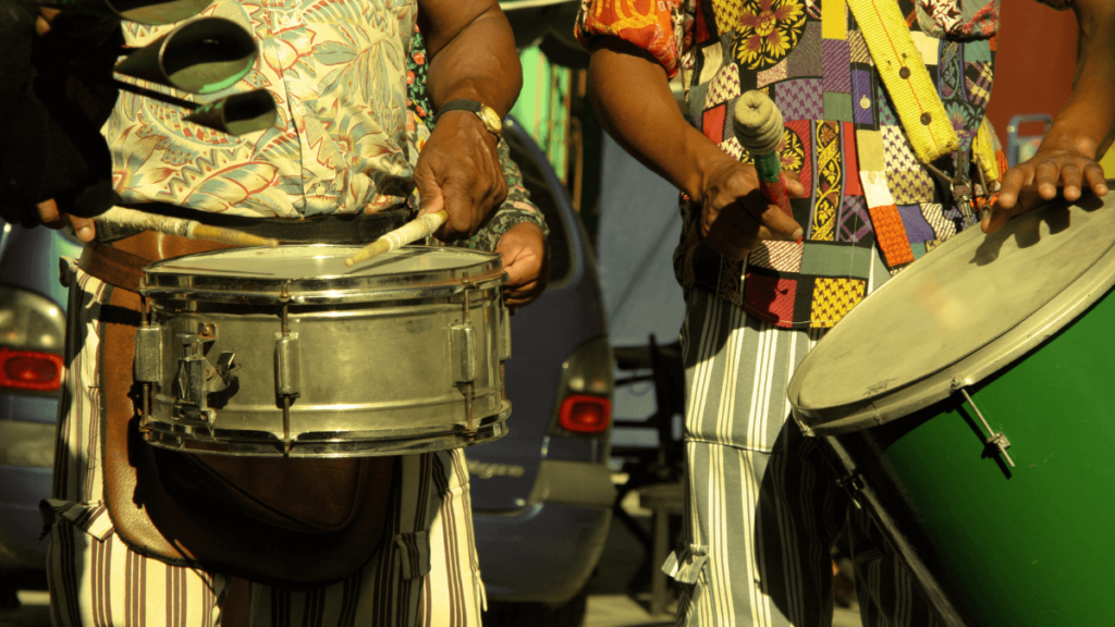 Close-up image of two people playing traditional samba drums, with a focus on their hands and the instruments, capturing the essence of rhythm in a musical performance.