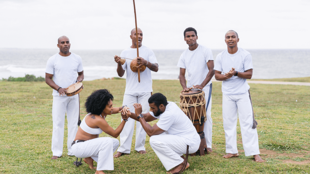 Group of people practicing capoeira on a grass field near the ocean, with some playing musical instruments while others engage in the martial art.