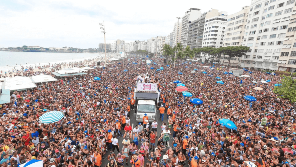 Aerial view of a massive carnival block party with a crowd surrounding a music truck on Rio de Janeiro's Copacabana beach.
