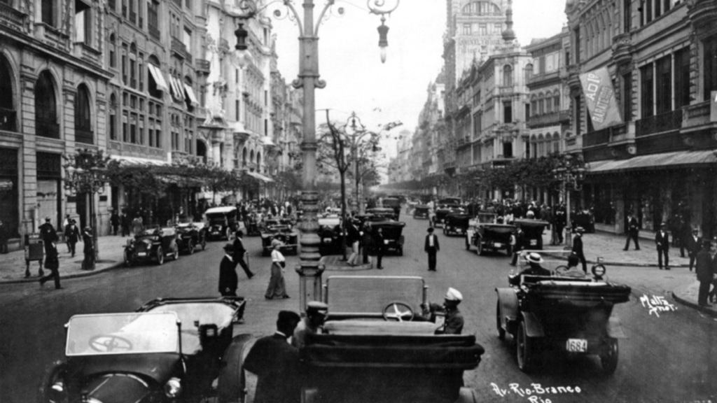 A busy street scene from early 20th century Rio de Janeiro, with vintage cars and people dressed in period attire.