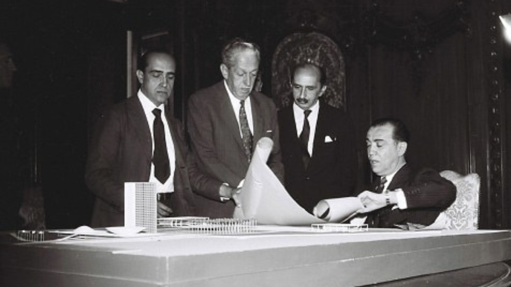 A black and white historical photo of four men reviewing architectural plans, with a model building in the foreground.
