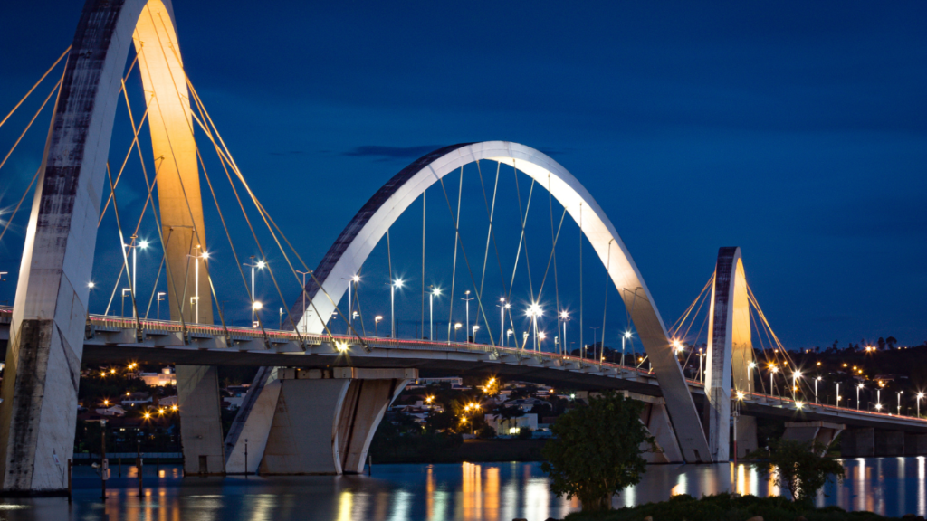 A night view of the Juscelino Kubitschek Bridge in Brasília, illuminated against the evening sky.