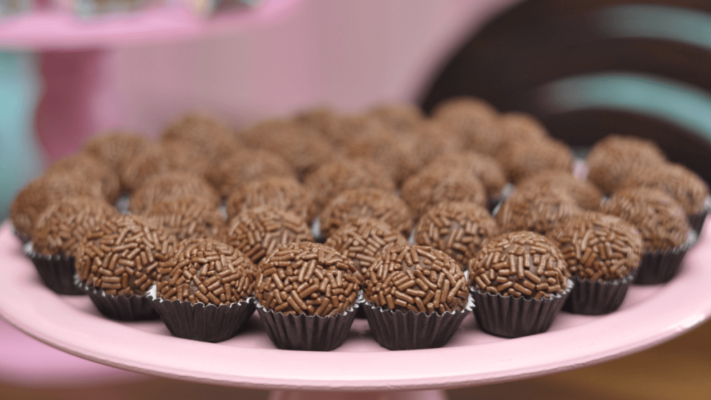 A spread of brigadeiros, Brazilian chocolate sweets, covered in chocolate sprinkles, placed in small paper cups on a pink plate, evoking a party dessert setting.