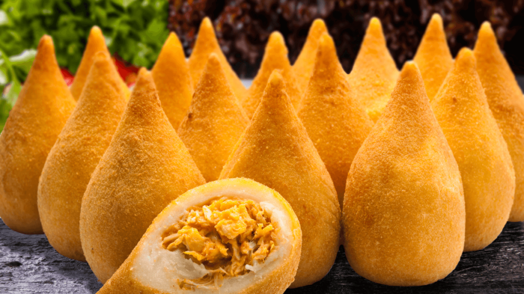 A coxinha, a popular food in Brazil, with a crispy golden exterior and stuffed with savory chicken filling, presented against a backdrop of fresh greens.