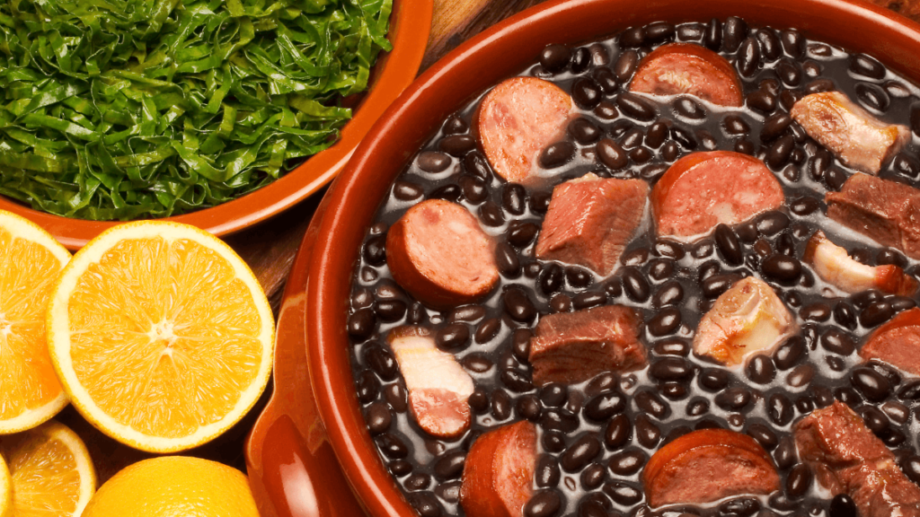 A traditional Brazilian feijoada, a stew of beans with beef and pork, in a terracotta pot, accompanied by chopped kale and orange slices on the side.