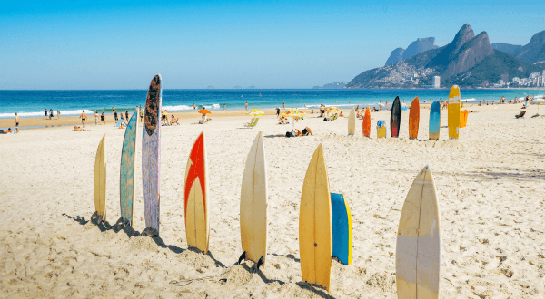 Colorful surfboards stand upright in the sand on Ipanema Beach, with a clear blue sky overhead and the iconic Two Brothers mountain in the background, capturing the vibrant spirit of Brazil's beach culture.