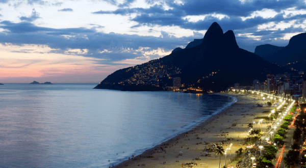 Twilight descends on Ipanema Beach as the city lights begin to twinkle, with the distinctive silhouette of the Dois Irmãos mountains standing guard over this bustling Brazil beach.