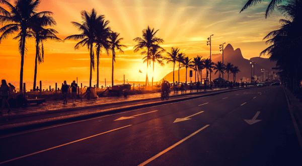 The sun sets behind the palm trees lining Ipanema Beach, creating a dramatic orange and purple sky, casting long shadows and highlighting the relaxed atmosphere of this famous Brazil beach.