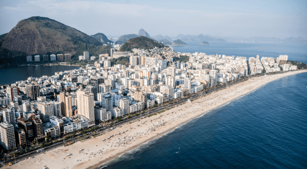 A distant view of Ipanema Beach set against the urban backdrop of Rio de Janeiro, with the calm blue ocean waters meeting the extensive sandy shores of this popular Brazil beach.