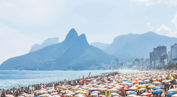 The crowded Ipanema Beach under a haze, with colorful umbrellas dotting the sand and the Dois Irmãos mountains rising in the background, a typical scene at this famous Brazil beach.