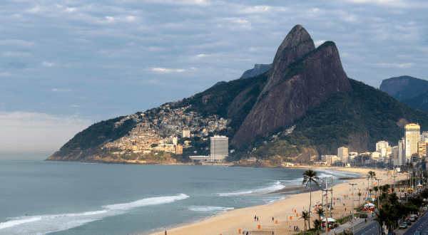 A tranquil view of Ipanema Beach from a high vantage point, showing the gentle curve of the shoreline, the calm ocean, and the cityscape of Rio de Janeiro beneath a cloudy sky.