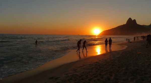 Silhouettes of people are seen on Ipanema Beach during a beautiful sunset, with the sun dipping below the horizon and casting a warm, golden light across the water and sand.