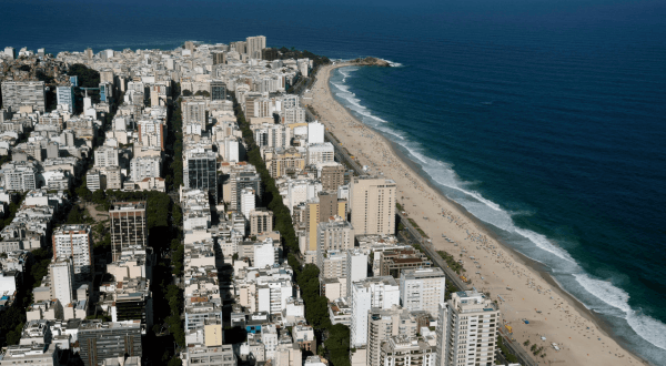 A bird's-eye view of Ipanema Beach showcases the dense urban layout of Rio de Janeiro, with the beach itself acting as a border between the city and the Atlantic Ocean, highlighting the contrast between nature and urbanization.