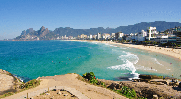 A view from the rocks overlooking the clear blue waters of Ipanema Beach, with the iconic Dois Irmãos peaks in the distance and a vibrant blue sky above Rio de Janeiro's famous Brazil beach.