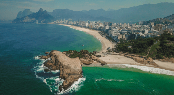 An aerial perspective of Ipanema Beach showing a rocky outcrop reaching into the sea, the long stretch of sand, and the dense urban landscape of Rio de Janeiro under a clear sky.