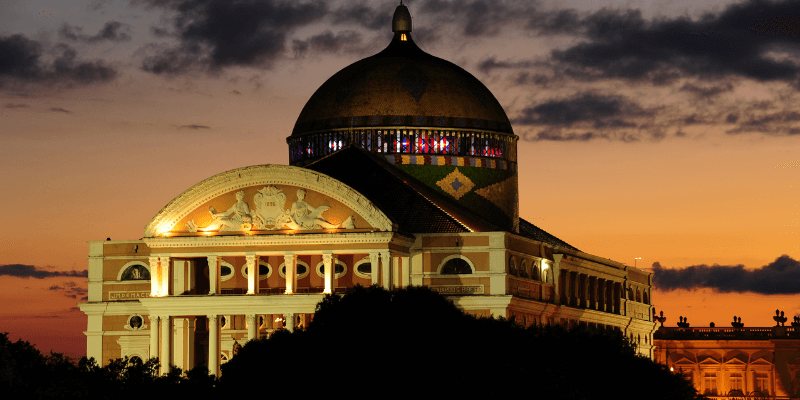 The historic Teatro Amazonas in Manaus at dusk, its illuminated dome and ornate architecture standing out against a purple and orange sky, symbolizing cultural richness in the Amazon.