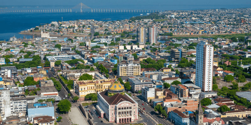 Aerial view of Manaus city with its mix of historic and modern buildings, including the famous Teatro Amazonas, set against the backdrop of the vast Amazon River.