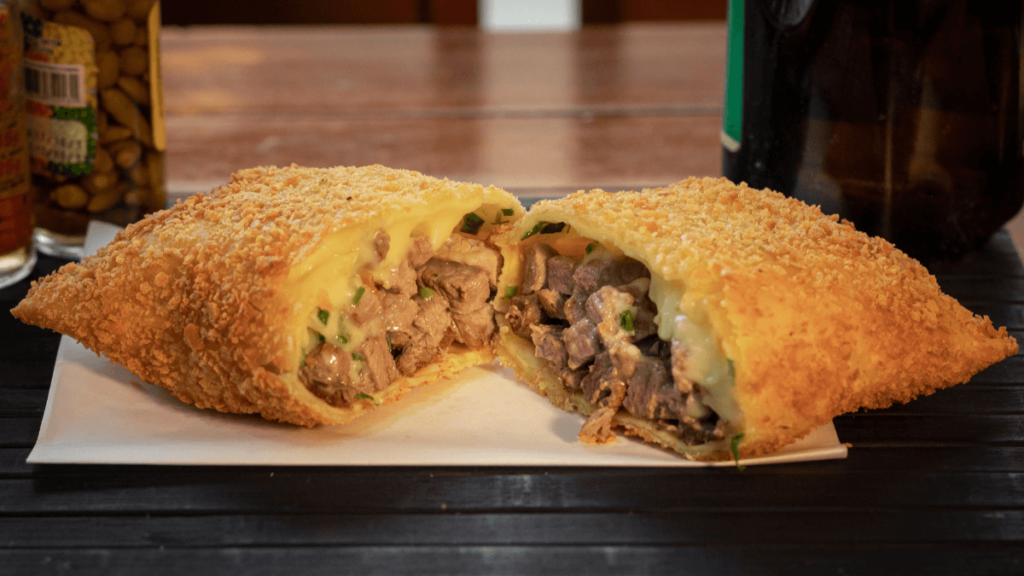 A pastel, a Brazilian stuffed pastry, sliced open to reveal a creamy filling of cheese and meat, presented on a simple white paper, evoking a street food vibe.