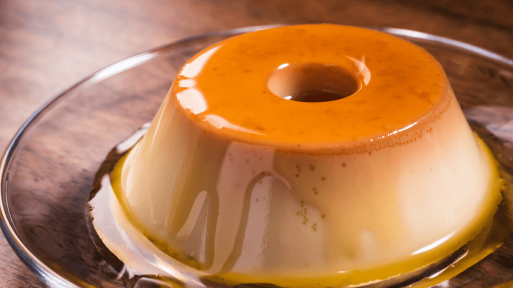 A caramel-topped pudim, a smooth Brazilian flan, on a glass serving plate, with a rich golden color and creamy texture highlighted by the lighting.