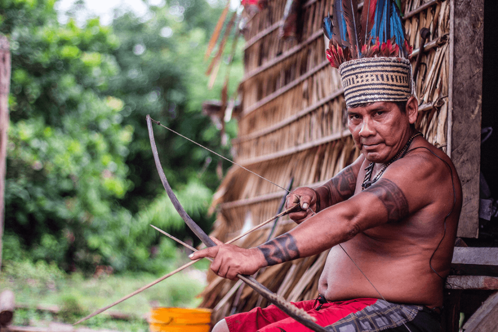 A Satére-Mawé man with a feathered headdress and facial tattoos aiming a bow and arrow, with a thatched hut in the background.