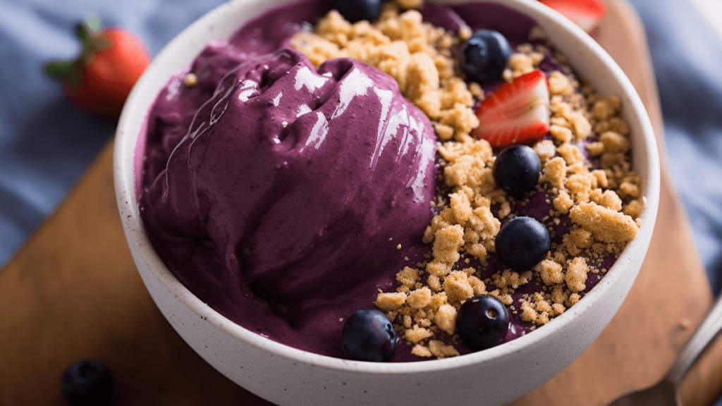 A bowl of açaí with a velvety purple texture, garnished with strawberries, blueberries, and granola, suggesting a fresh and healthy snack.