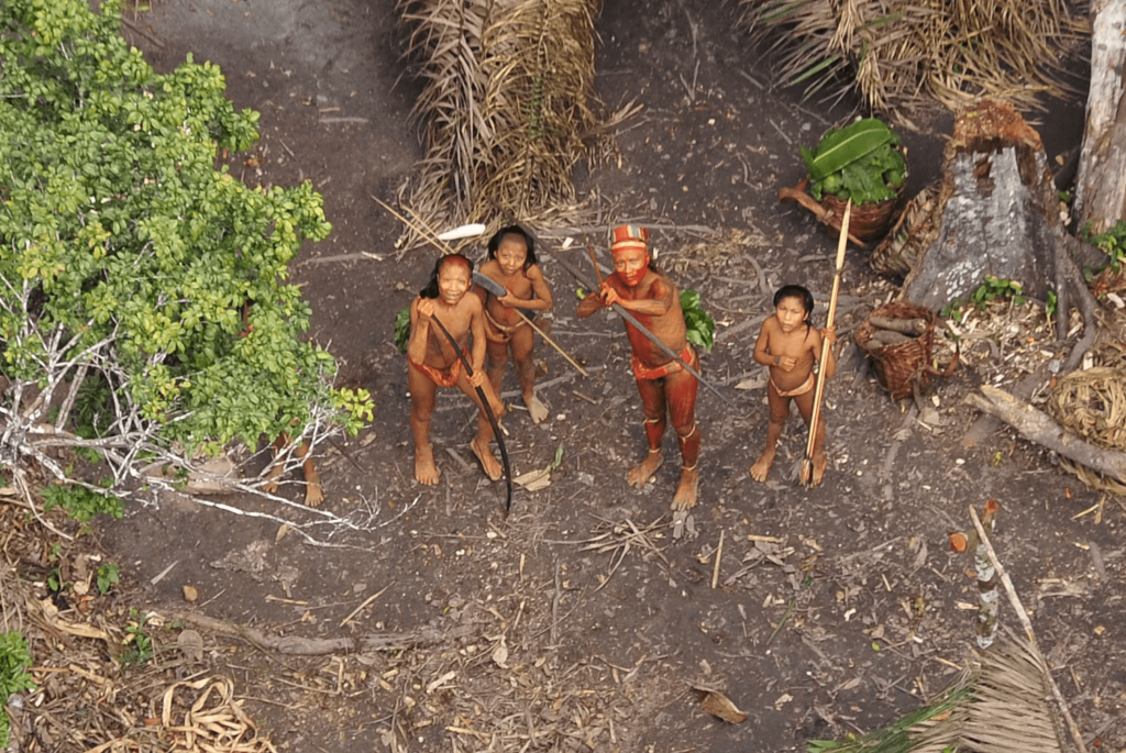 Alt Text: "An aerial view of a small group of indigenous people from an isolated tribe in the Amazon rainforest. The group, consisting of adults and children, stands barefoot amidst their forest environment. They are clad in traditional red body paint and minimal clothing, holding bows and looking up, possibly at a drone or camera. Surrounding them are natural elements of their habitat, including green foliage, a thatched structure, and everyday tools, showcasing a glimpse of their self-sustained lifestyle in harmony with nature.