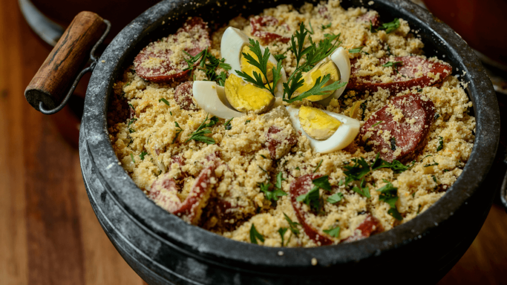 Farofa is garnished with slices of sausage and boiled eggs, and it's all presented in a black pot with a wooden handle, accompanied by fresh parsley, creating a festive and authentic meal presentation.