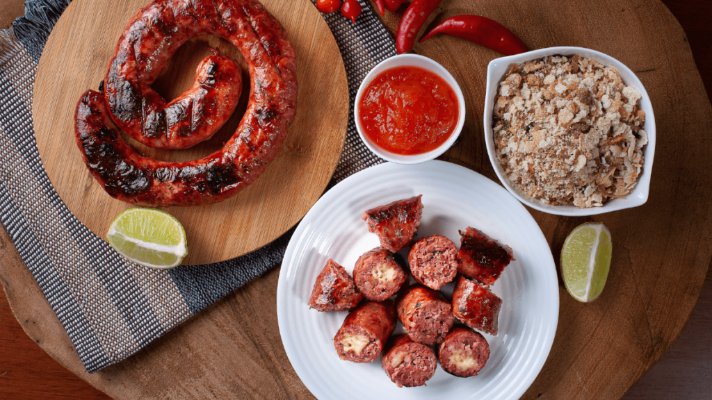 This image displays a wooden cutting board with a large, coiled grilled sausage on it, accompanied by a small bowl of red chili sauce and a bowl of crumbled, seasoned cassava flour mixture known as farofa. There are also two lime wedges and some fresh red chilies on the side. The plate of sliced sausage shows the juicy interior, hinting at a spicy and savory flavor profile.