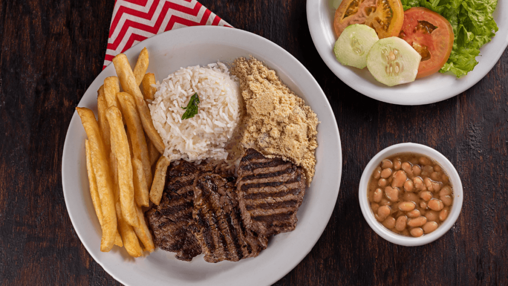 The photo presents a traditional Brazilian meal with a grilled steak, accompanied by sides of golden French fries, white rice, a scoop of farofa, fresh salad consisting of lettuce, tomato, and cucumber slices, and a small bowl of cooked beans. The dish is served on a white plate with a red and white striped napkin on the side, suggesting a hearty and flavorful dining experience.