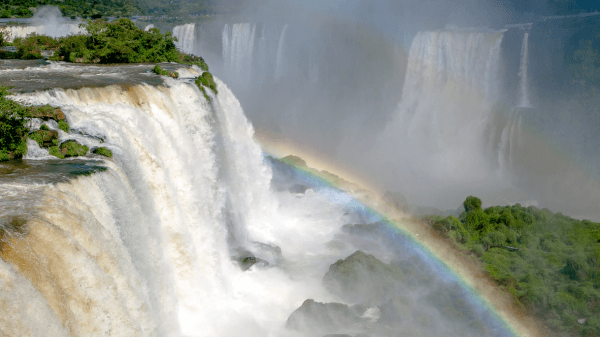 A vibrant rainbow arcs over the powerful Iguazu Falls surrounded by lush greenery, as mist rises from the thundering waters.
