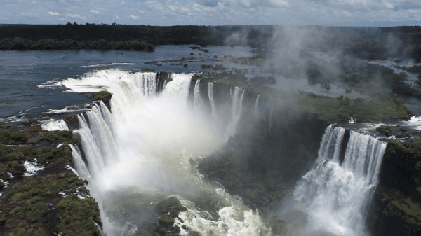 The misty atmosphere above Iguazu Falls in Brazil, with cascades flowing powerfully into a vast river surrounded by dense forest.