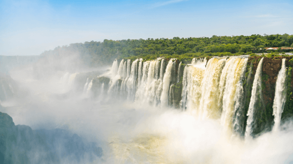 The sun casts a bright haze over the extensive Iguazu Falls, highlighting the massive scale and majestic beauty of the cascades.