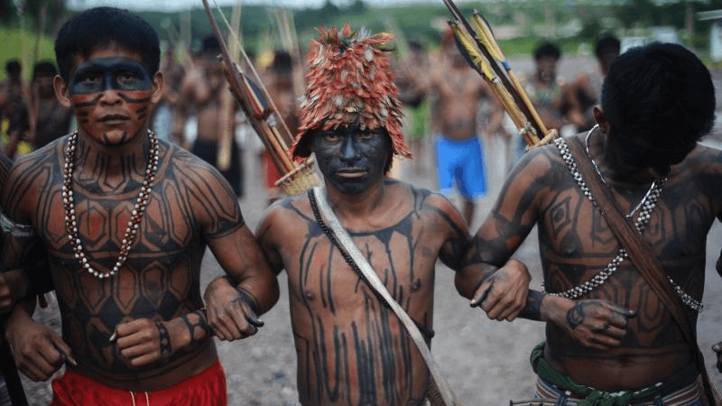 Three Munduruku men with traditional body paint and feather headdresses holding long spears, standing confidently in a village setting.
