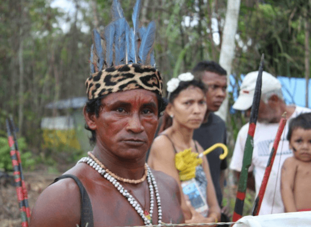 A Mura man in a jaguar-patterned headband and blue feather headdress, with a contemplative expression, stands before other community members.