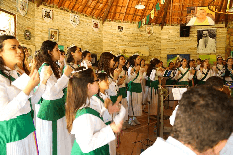 A choir dressed in white and green robes clapping and singing in a Santo Daime ceremony, with religious iconography in the background.