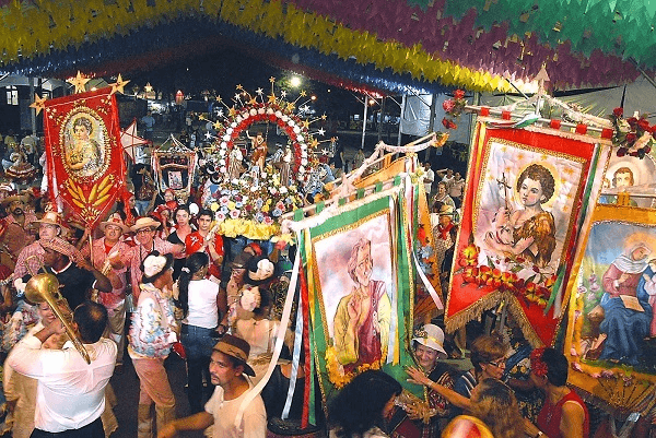 A festive scene from a Brazilian cultural religious festival, with people carrying ornate banners and images of saints, indicative of syncretic celebrations blending Catholic and local traditions.