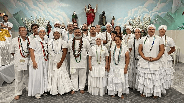 A group of people dressed in white ceremonial clothes posing in front of religious statues and images, representing practitioners of Umbanda, Afro-Brazilian religion.