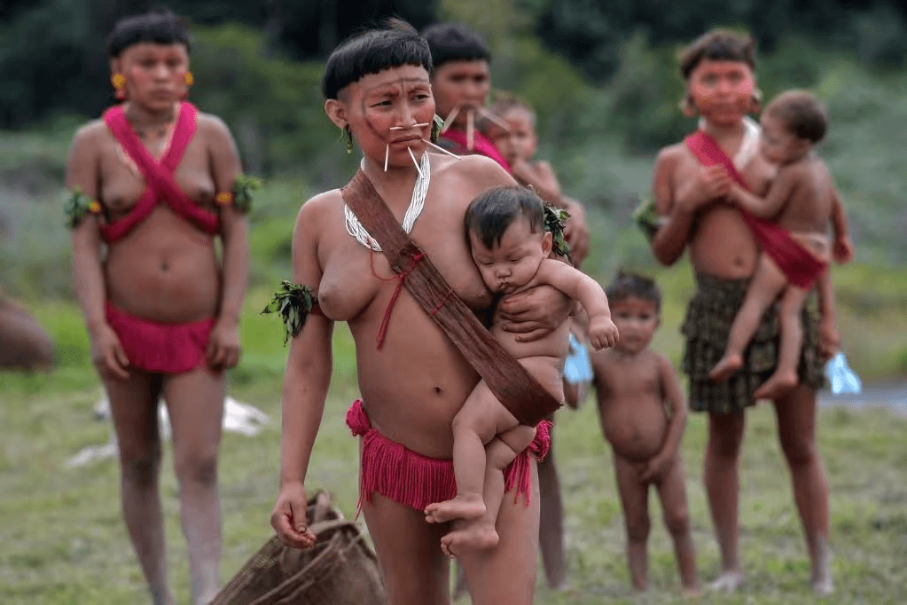 A Yanomami man in ceremonial attire carries a baby, with children in the background, highlighting family and tribal life.