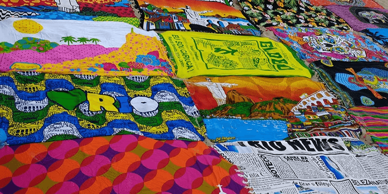 A colorful display of assorted textiles featuring various designs, with prominent "Rio" lettering, reflecting Brazilian culture and landmarks, laid out for sale.