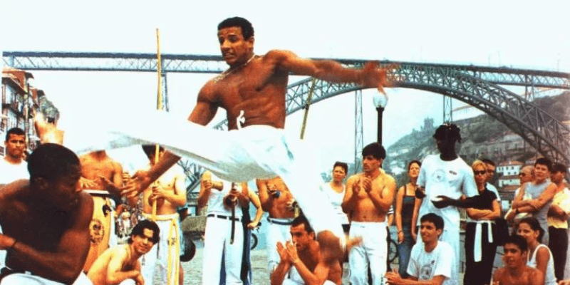 An action shot of two Brazilian Capoeira practitioners sparring in front of an audience, with one executing a high kick, near an iconic bridge structure, illustrating the cultural heritage of the martial art.