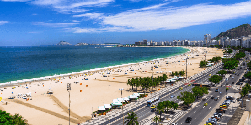 A scenic view of Copacabana, one of the most famous beaches in Rio de Janeiro beach with a distinctive wavy sidewalk pattern, crowded with people enjoying the sun, and city traffic moving alongside the beachfront.