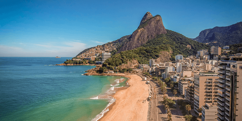 A scenic coastal view showing a curving sandy beach next to a high mountain peak, with residential buildings nestled between the greenery and the shoreline under a clear blue sky.