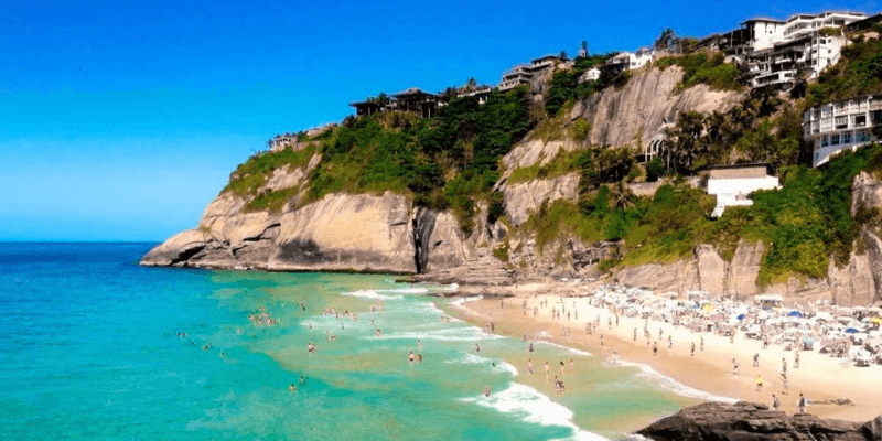 A vibrant beach scene with people swimming and sunbathing, set against a backdrop of a steep cliff with lush vegetation and luxury houses perched on the edge, overlooking the clear blue water.