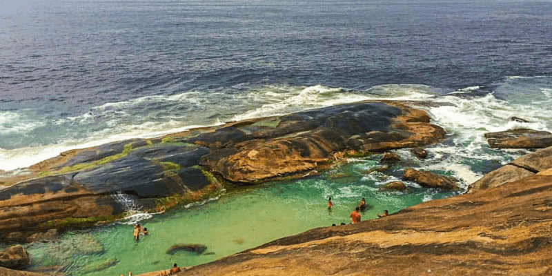 A natural tide pool on coastal rock formations, with people enjoying the water, set against the rough sea and a hazy horizon.