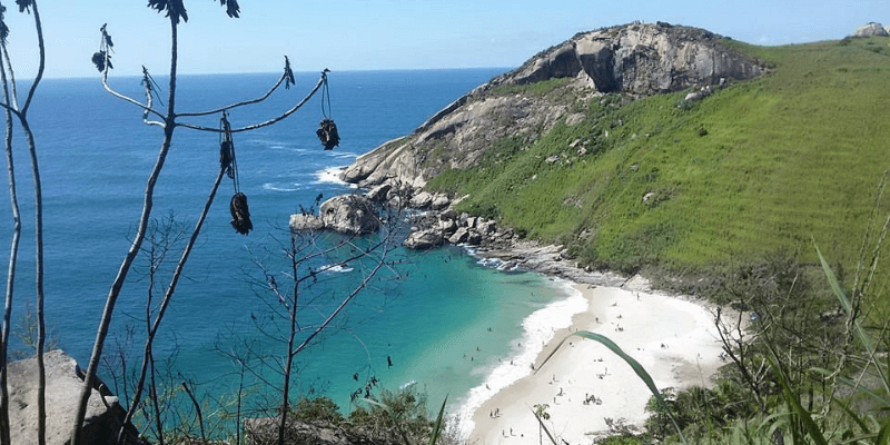A hidden cove with a secluded white sand beach surrounded by green hills and rocky outcrops, with clear turquoise water inviting a relaxing swim.