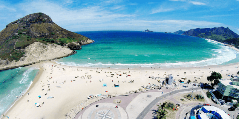 A bird's-eye view of a bustling beach with a circular plaza, beachgoers enjoying the sun, clear turquoise waters, and a mountainous landscape in the distance.