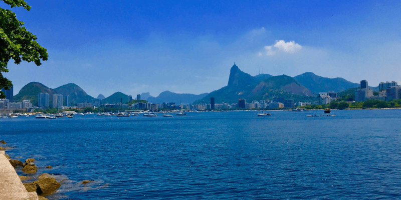 A picturesque view of a bay with numerous boats, a cityscape in the background, and the iconic Christ the Redeemer statue visible atop a distant mountain under a clear blue sky.