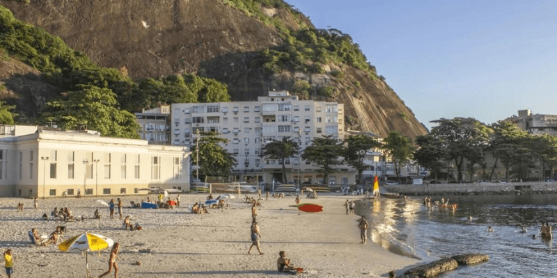 A sunny beach scene with people enjoying the waterfront, the cityscape close to the shore, and a mountain in the background, creating a harmonious blend of urban and natural elements.